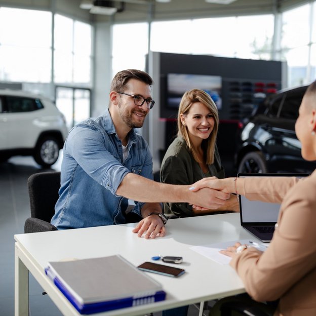 Zwei Personen sizten im Autohaus vor einem Schreibtisch eines Mitarbeiter und schütteln ihm die Hand