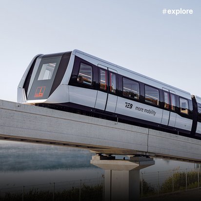 Eine moderne Einschienenbahn mit der Aufschrift "more mobility" fährt über eine erhöhte Betontrasse, die sich über Wasser und eine bewaldete Landschaft erstreckt. Der silber-graue Zug spiegelt sich im ruhigen Wasser darunter wider.