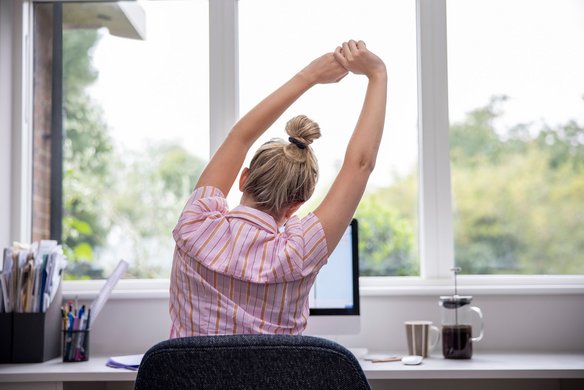 Person streckt sich am Schreibtisch vor einem Fenster während der Arbeit im Homeoffice.