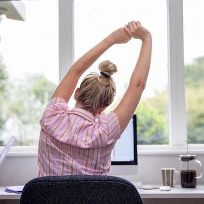 Person streckt sich am Schreibtisch vor einem Fenster während der Arbeit im Homeoffice.