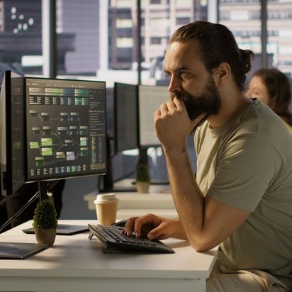 Datenanalyse im Fokus – Programmierer bei der Arbeit Mann mit Bart arbeitet fokussiert am Computer mit Datenvisualisierung auf dem Monitor im modernen Büro