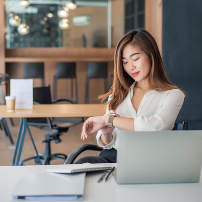 Frau am Schreibtisch schaut auf ihre Smartwatch, Laptop und Notizbuch vor sich