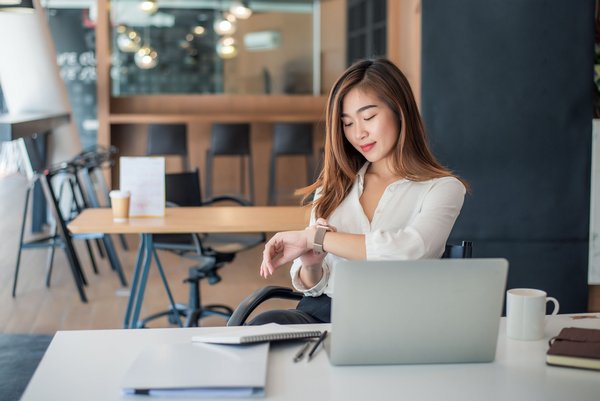 Frau am Schreibtisch schaut auf ihre Smartwatch, Laptop und Notizbuch vor sich