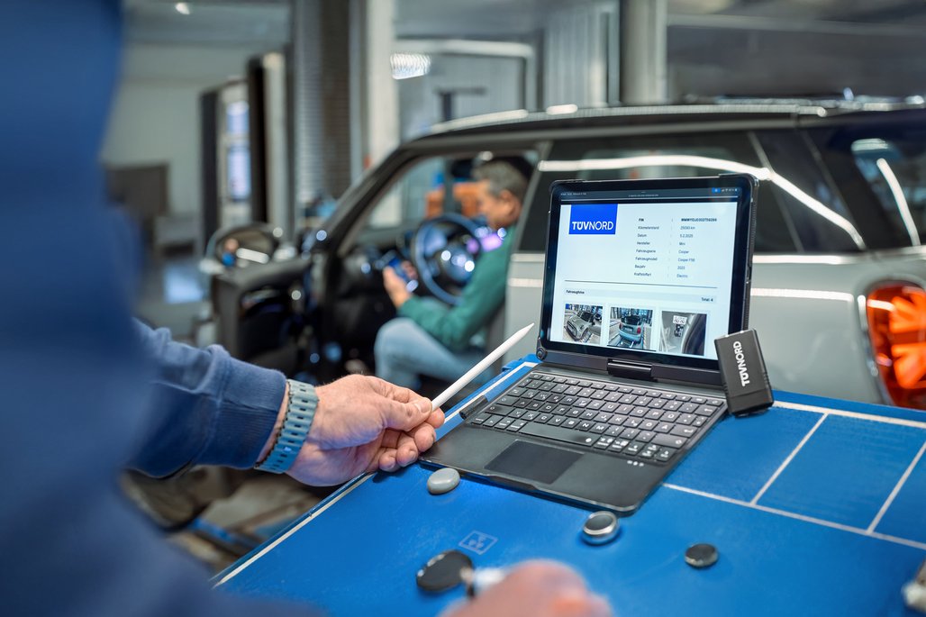 Ein Tablet mit TÜV Nord Fahrzeugbericht eines Mini Cooper steht in einer Werkstatt auf einem Tisch, im Hintergrund sitzt eine Person im Fahrzeug.