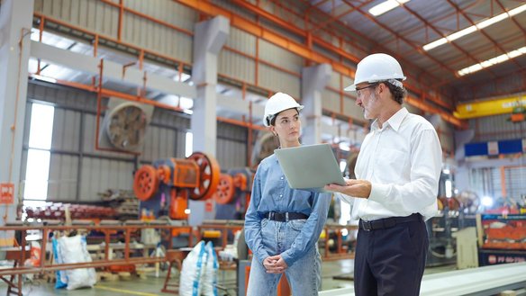 Zwei Personen mit Schutzhelmen im Gespräch in einer Industriehalle, einer hält einen Laptop