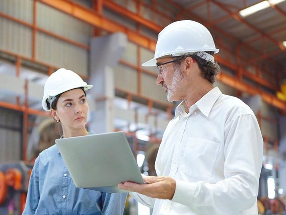 Zwei Personen mit Schutzhelmen im Gespräch in einer Industriehalle, einer hält einen Laptop