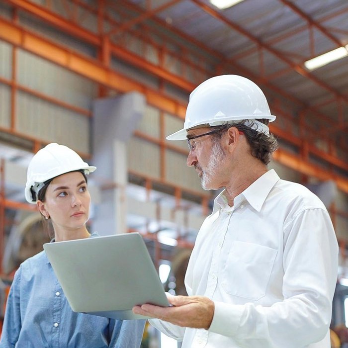 Zwei Personen mit Schutzhelmen im Gespräch in einer Industriehalle, einer hält einen Laptop
