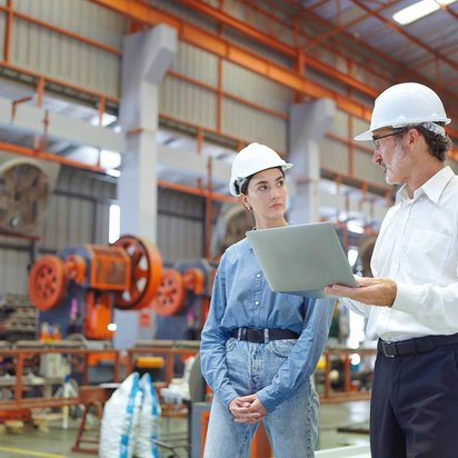 Technikgespräch in der Produktionshalle mit digitaler Unterstützung Zwei Personen mit Schutzhelmen im Gespräch in einer Industriehalle, einer hält einen Laptop