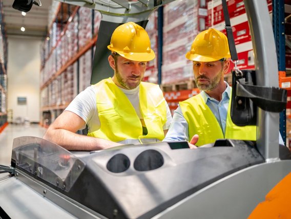 Tuev Nord Gabelstapler Ausbildung Zwei Personen in gelben Sicherheitswesten und Schutzhelmen stehen in einem Lagerhaus neben einem Gabelstapler. Sie sind in ein Gespräch vertieft, während sie die Umgebung im Lager betrachten.