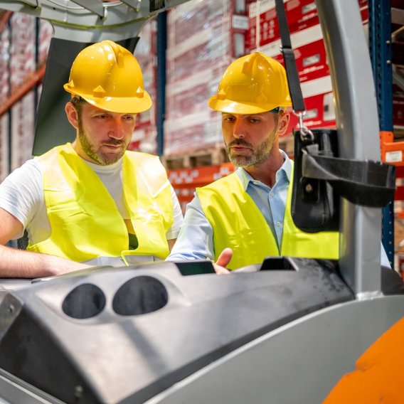  Zwei Personen in gelben Sicherheitswesten und Schutzhelmen stehen in einem Lagerhaus neben einem Gabelstapler. Sie sind in ein Gespräch vertieft, während sie die Umgebung im Lager betrachten.