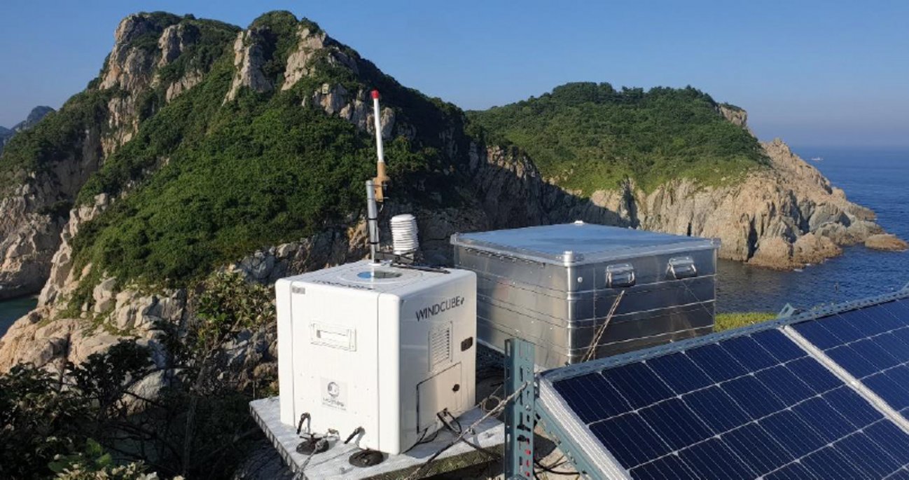 Eine Messstation mit weißen Geräten und blauen Solarpanels steht auf einer Klippe vor dramatischer Küstenlandschaft mit bewaldeten Felseninseln und tiefblauem Meer. Die wissenschaftliche Ausrüstung wird durch nachhaltige Solarenergie betrieben und überwacht Umweltdaten in abgelegener Meereslage.