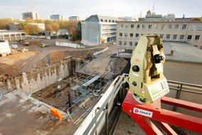  Eine Baustelle in einer städtischen Umgebung, auf der ein großes Baugrubenloch zu sehen ist, umgeben von Baugerüsten und Erdbewegungsmaschinen. Im Vordergrund steht ein modernes Vermessungsgerät auf einem roten Stativ mit dem Logo "DMT". Im Hintergrund sind mehrere Gebäude und Kräne zu erkennen, die auf eine aktive Bauphase hinweisen. Die Umgebung ist von Bäumen und weiteren städtischen Strukturen umgeben.