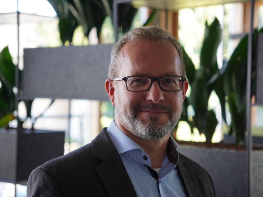 Michael Baranowski mit Brille und Bart trägt einen dunklen Blazer und steht in einem Büro mit Pflanzen im Hintergrund.