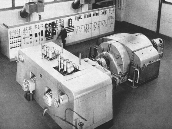 Schwarz-weiß-Aufnahme einer industriellen Dampfturbine mit Kontrollpult in einem Kraftwerksraum der 1950er oder 1960er Jahre.