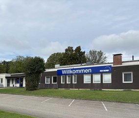 TÜV NORD Station Iserlohn mit großem „Willkommen“-Schild, Grünflächen und Parkplatz, Bäume im Hintergrund.