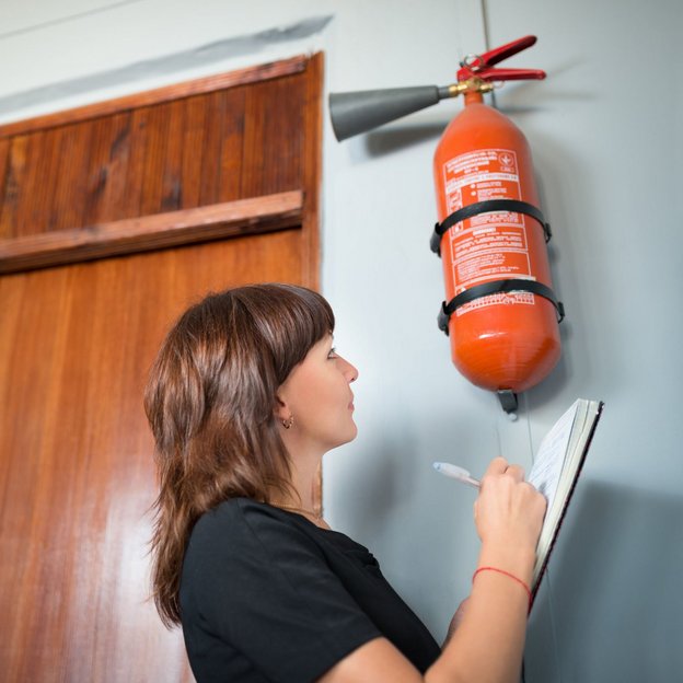 Frau überprüft Feuerlöscher an der Wand und notiert Daten in ein Klemmbrett