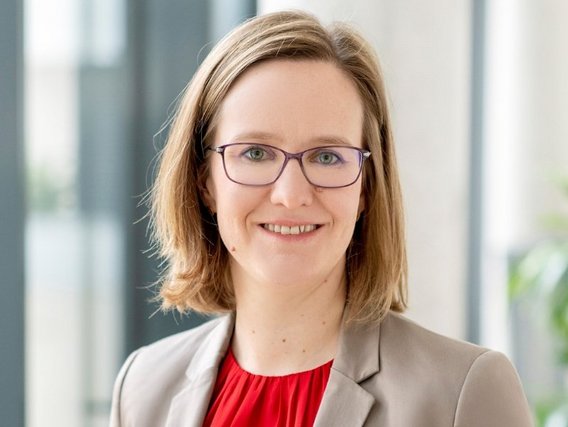 Susanne Günzerodt mit schulterlangem blondem Haar und lila Brille trägt einen beigen Blazer mit roter Bluse und lächelt in die Kamera vor einem hellen Hintergrund.