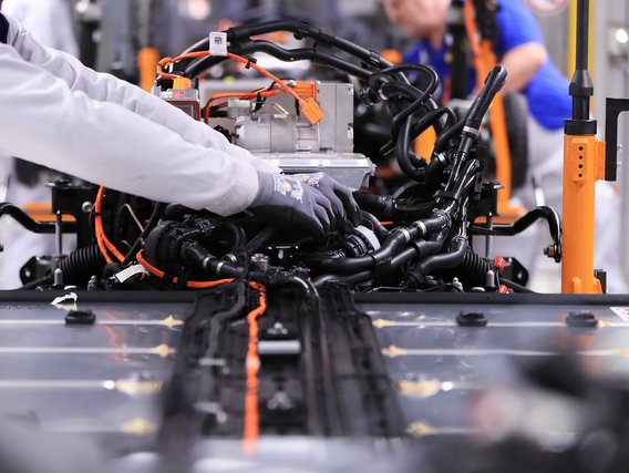 Eine Person in weißem Laborkittel mit grauen Schutzhandschuhen montiert Kabel und Komponenten an einem Elektroauto-Antriebssystem mit schwarzen Kabeln und orangefarbenen Verbindungselementen auf einer Fertigungslinie.