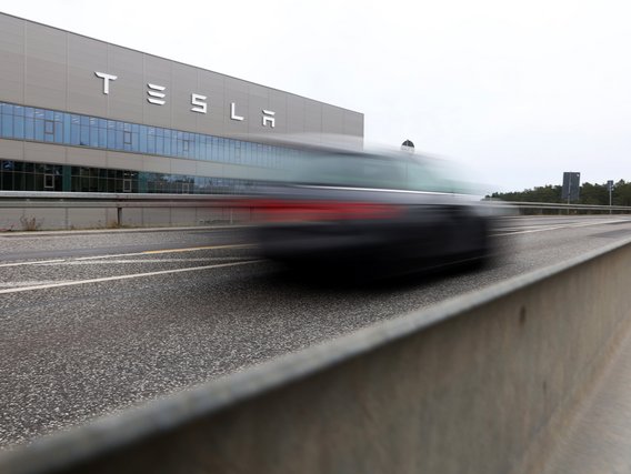 Ein verschwommenes Auto fährt an einem Tesla-Gebäude vorbei, das mit dem Markenlogo an der grauen Fassade und blauen Glasfenstern zu sehen ist.