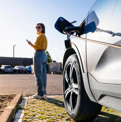 TÜV NORD-Ladesäule Frau steht neben einem Elektroauto an einer Ladestation und nutzt ihr Smartphone.