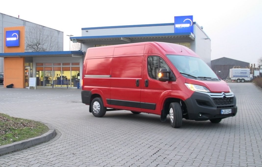 Ein roter Kastenwagen steht vor einem modernen Autohaus mit blau-oranger Fassade auf gepflastertem Vorplatz. Das Nutzfahrzeug zeigt die praktische Seite nachhaltiger Mobilität für gewerbliche Zwecke.