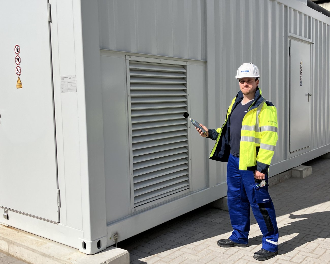Eine Person in Sicherheitskleidung und Helm führt eine Inspektion an einem großen weißen Container durch. Sie hält ein Messgerät in der Hand. Der Container hat Lüftungsschlitze und Warnschilder auf der Tür.