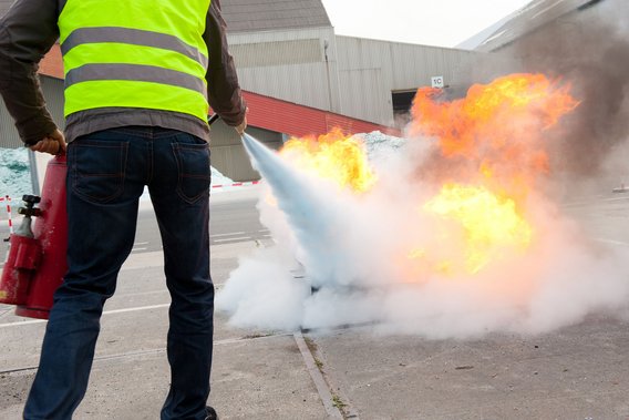 Person mit Warnweste löscht bei einer Übung im Freien ein offenes Feuer mit einem Feuerlöscher.