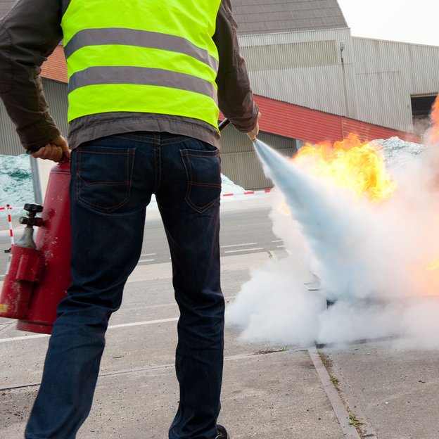 Person mit Warnweste löscht bei einer Übung im Freien ein offenes Feuer mit einem Feuerlöscher.