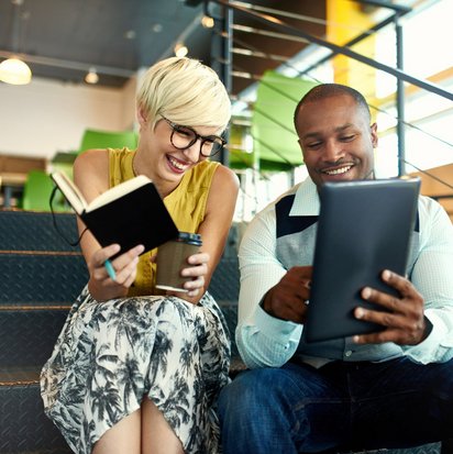 Zwei lächelnde Kollegen sitzen auf einer Treppe und arbeiten gemeinsam mit Tablet und Notizbuch.