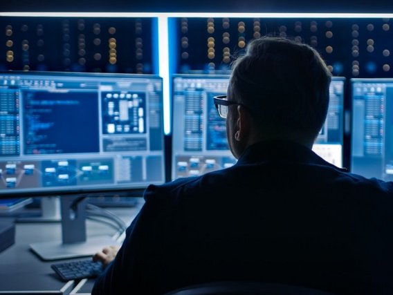 Eine Person sitzt vor mehreren Monitoren mit Code und Daten in einem dunklen Raum mit blinkenden Lichtern im Hintergrund.