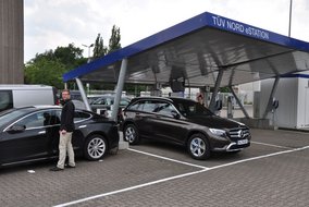 Eine TÜV NORD eStation zeigt Elektrofahrzeuge beim Ladevorgang unter einem modernen Überdach mit blauen Akzenten. Mehrere Personen sind bei der Nutzung der Ladeinfrastruktur zu sehen.