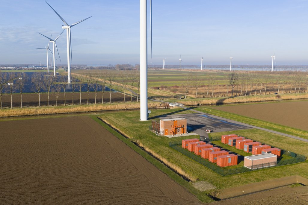 Eine Landschaft mit mehreren Windkraftanlagen und einem Energiespeicherbereich aus roten Containern. Im Hintergrund sind Felder und ein klarer blauer Himmel zu sehen. Die Windräder stehen in einer Reihe und erzeugen erneuerbare Energie.