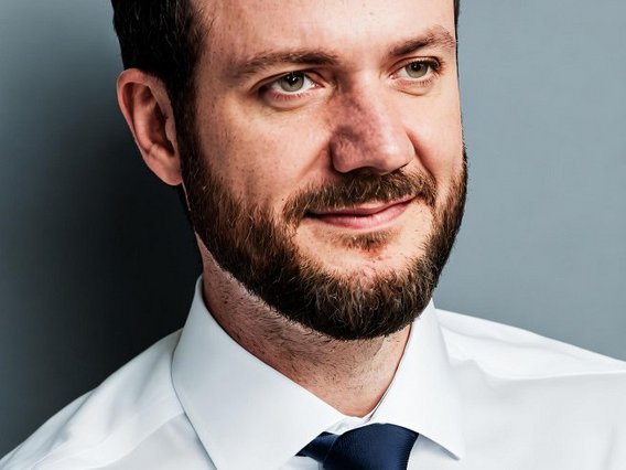 Nahaufnahme von Dr. Timo Bartkewitz in weißem Hemd und blauer Krawatte vor einem grauen Hintergrund.
