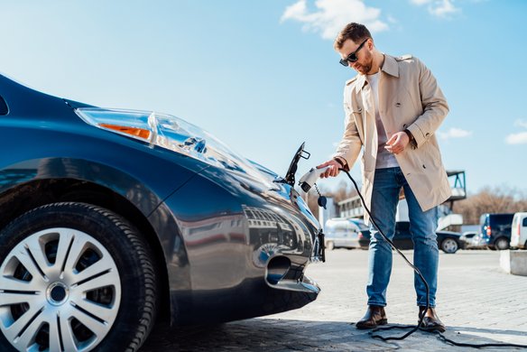 Mann im Trenchcoat steht mit einem Ladekabel in der Hand vor seinem Auto
