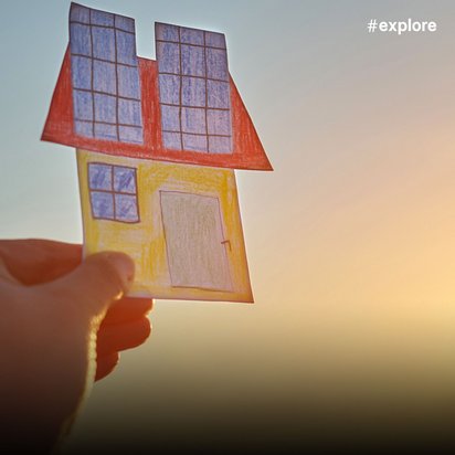 Eine Hand hält ein kleines Papierhaus mit rotem Dach, gelben Wänden und Solarpanels gegen einen warmen Sonnenuntergang. Das Miniaturhaus symbolisiert nachhaltiges Wohnen und erneuerbare Energie vor dem dramatischen Himmel mit goldenen Lichtstrahlen.