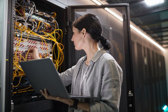 Technikerin arbeitet mit Laptop an einem offenen Serverschrank voller Kabel im Rechenzentrum.