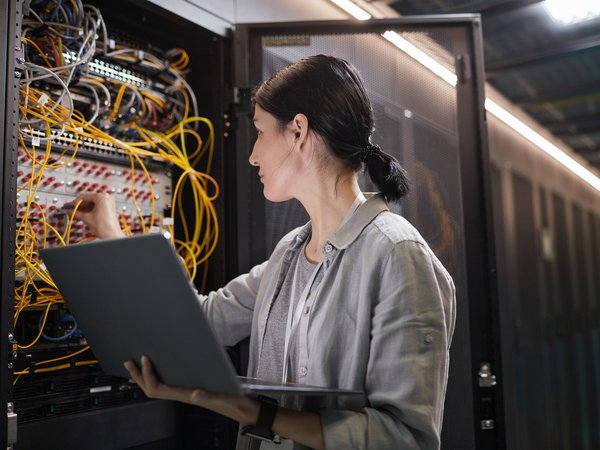 Technikerin arbeitet mit Laptop an einem offenen Serverschrank voller Kabel im Rechenzentrum.