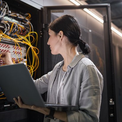 Technikerin arbeitet mit Laptop an einem offenen Serverschrank voller Kabel im Rechenzentrum.